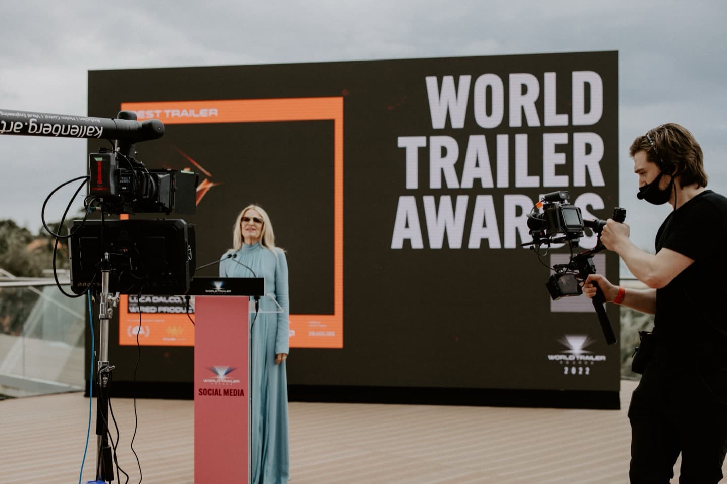 The World Trailer Awards stage — the presenter at a branded lectern, a camera on sticks and a gimbal operator covering her