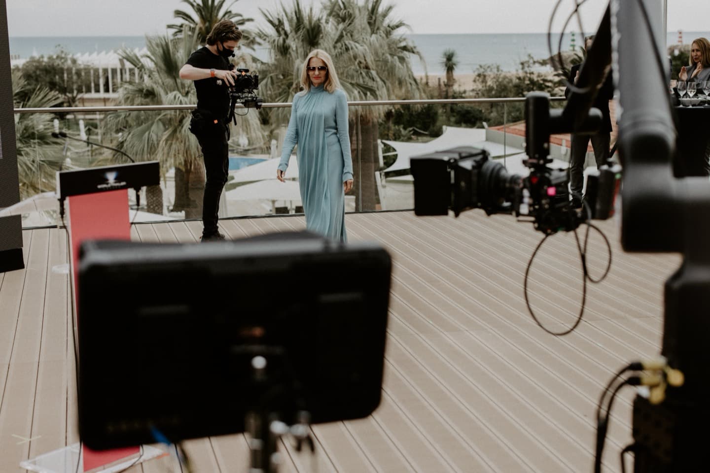 A terrace set-up above the sea: red carpet, camera and monitor in the foreground, the presenter walking the mark
