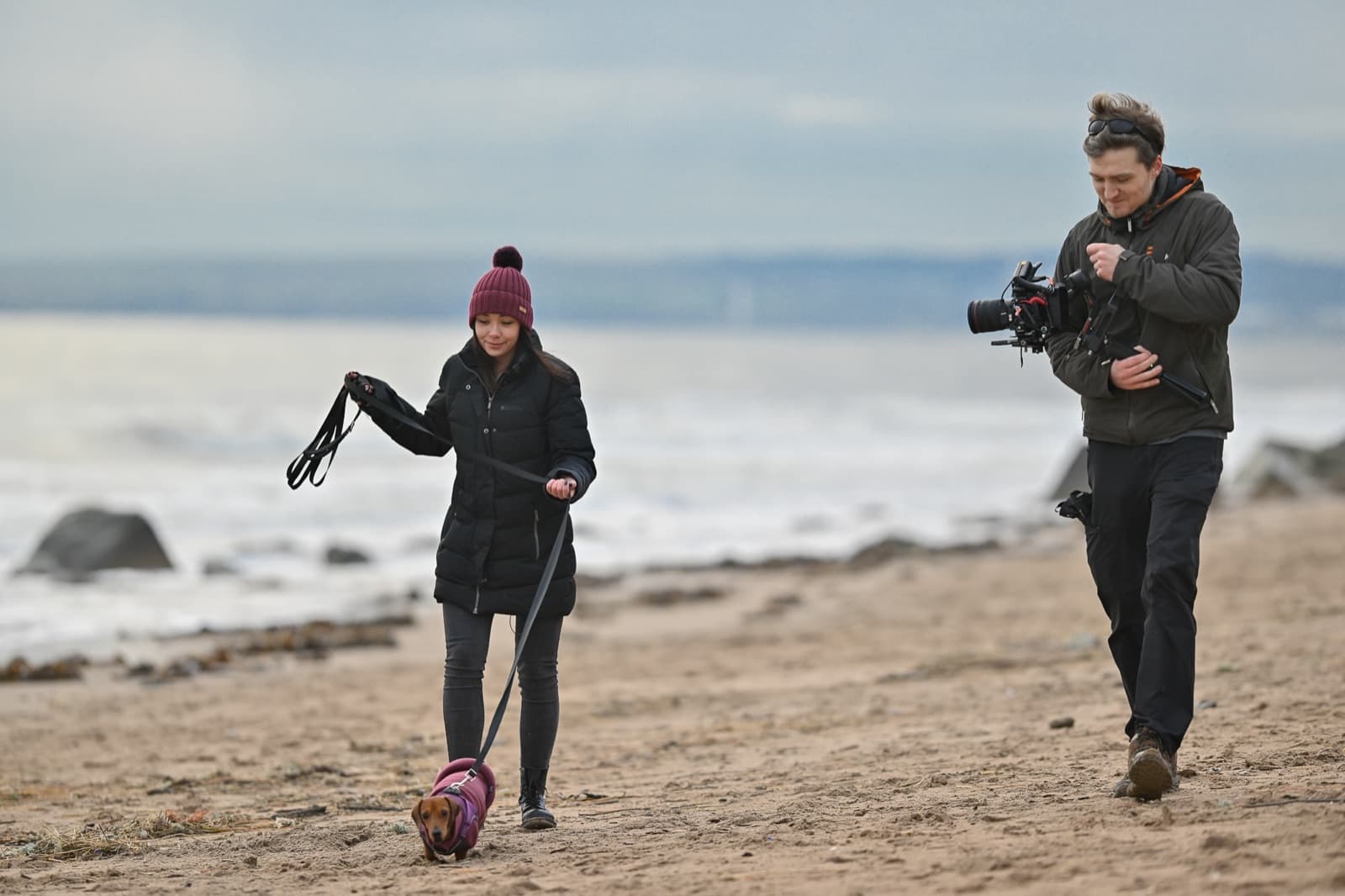 Tracking alongside a contributor walking a dog on a beach, the operator matching her pace with the camera rigged