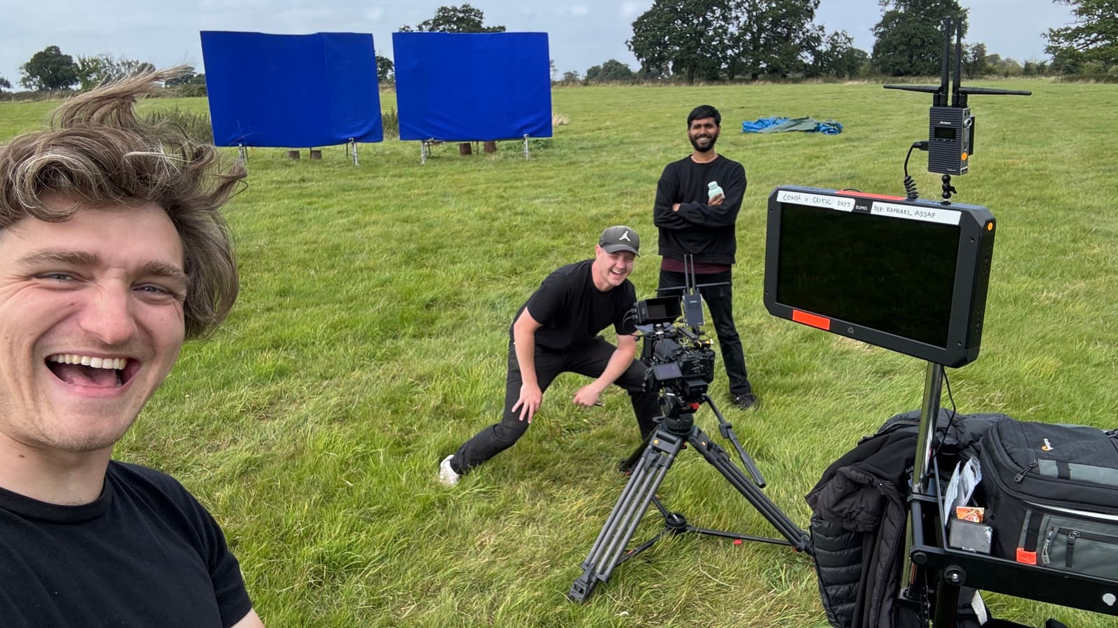 Two blue screens rigged out in an open field, the crew setting up around the camera