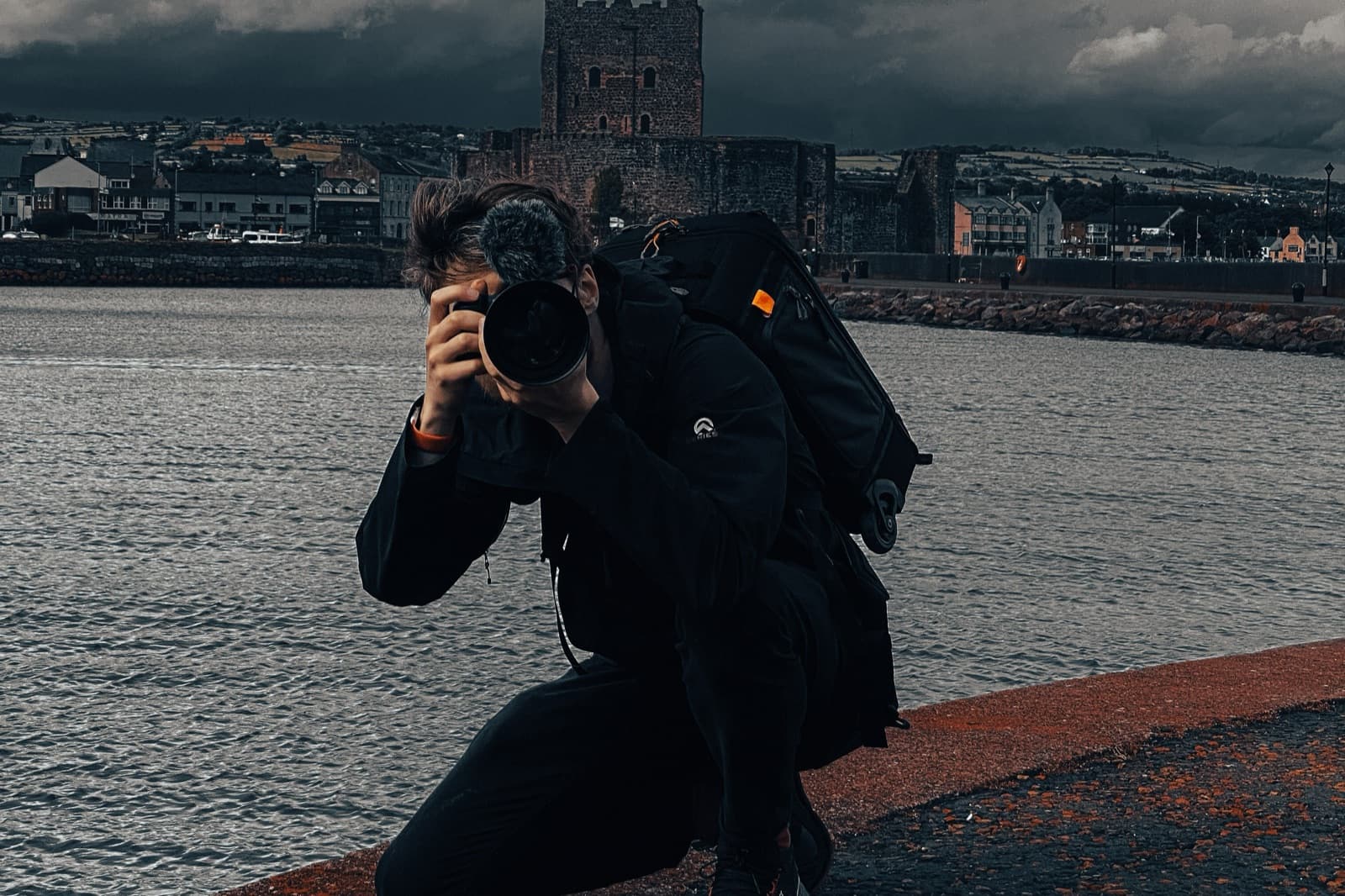 A camera operator kneeling on a sea wall for a low angle, castle and heavy cloud across the water behind