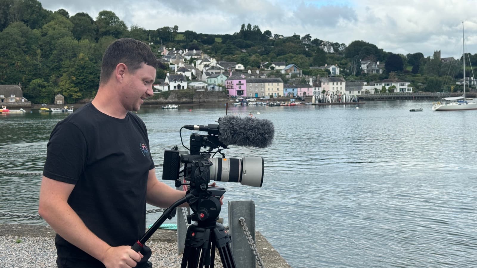 A camera operator setting up over a harbour, a town on the hillside across the water