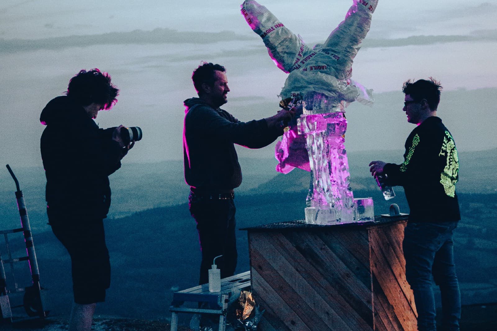 Filming an ice sculpture lit magenta on a hilltop at dusk, three crew around it