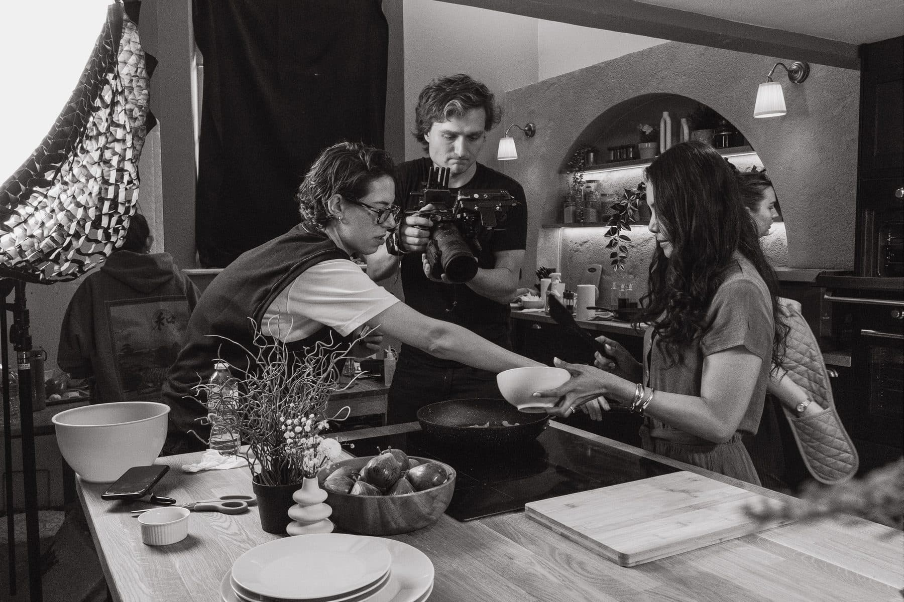 A kitchen set mid-take, camera operator close in on a hand-off between two presenters