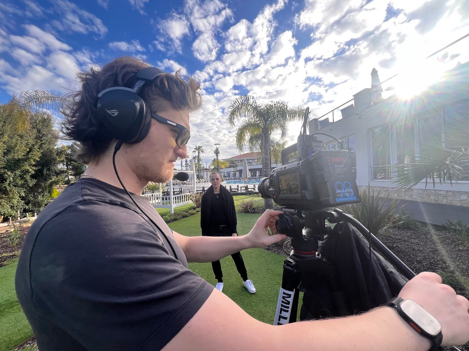 A camera operator on headphones setting up in strong sun, the subject waiting in the background