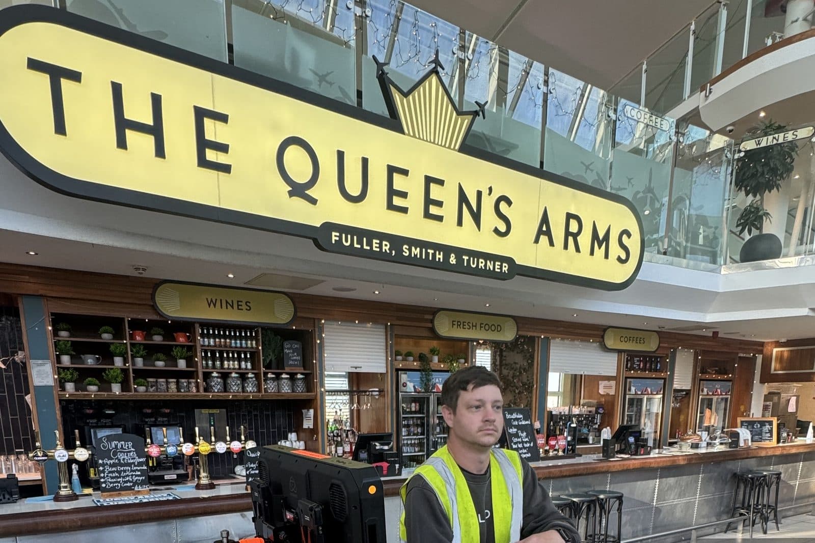 A loaded kit cart parked under a pub sign inside an airport terminal, one of the crew in a hi-vis vest waiting beside it