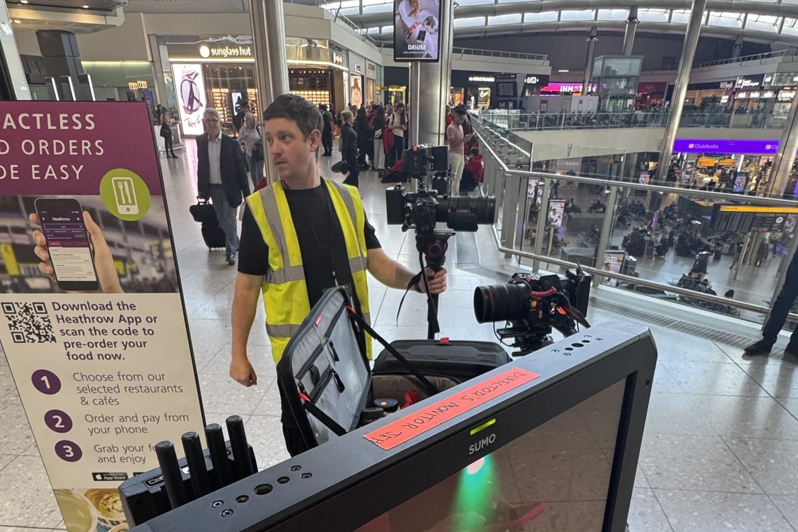 A crew member in a hi-vis vest carrying a rigged camera through a busy airport concourse, the monitor still up on the kit cart in the foreground