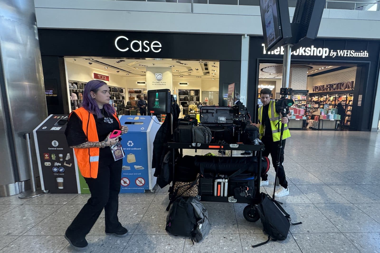 Two of the crew in hi-vis moving a loaded kit cart past the shops in an airport terminal