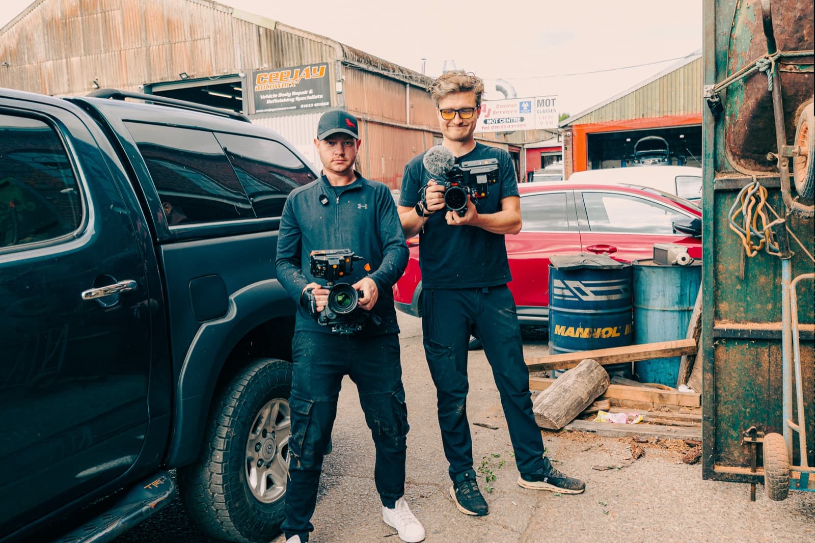 Two of the crew in a working yard beside a pickup, cameras built and ready between set-ups