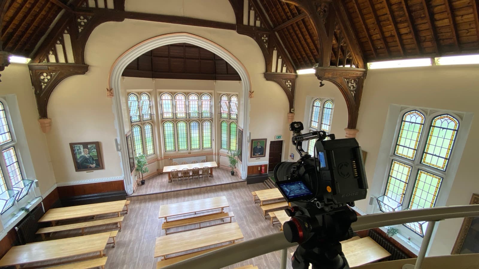 A camera set up in a timber-roofed hall under a gothic window, the benches cleared for the shoot