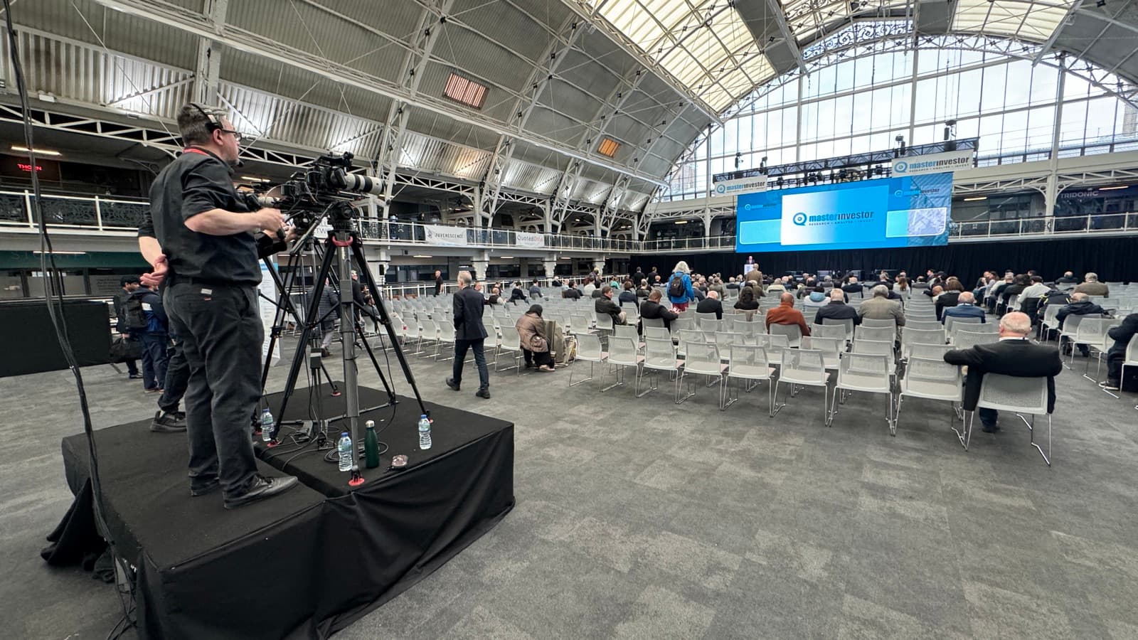 Two operators working a long lens from a camera riser at the back of a glass-roofed hall