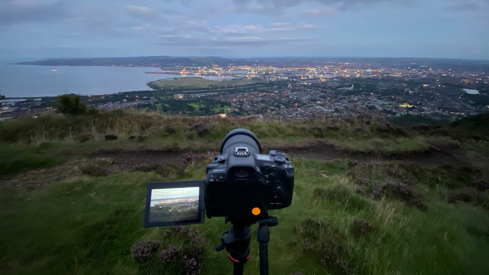 A camera locked off on a heather hilltop above a city at dusk, the frame live on the monitor