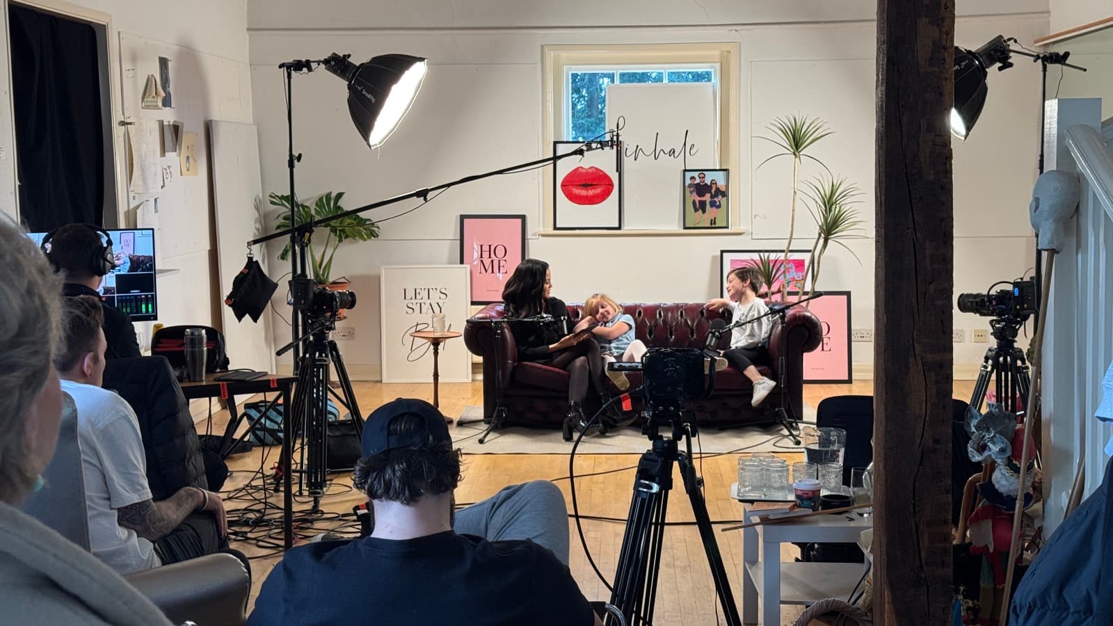 A loft studio mid-take, two contributors on a sofa under a boomed lamp and the room watching from behind the camera