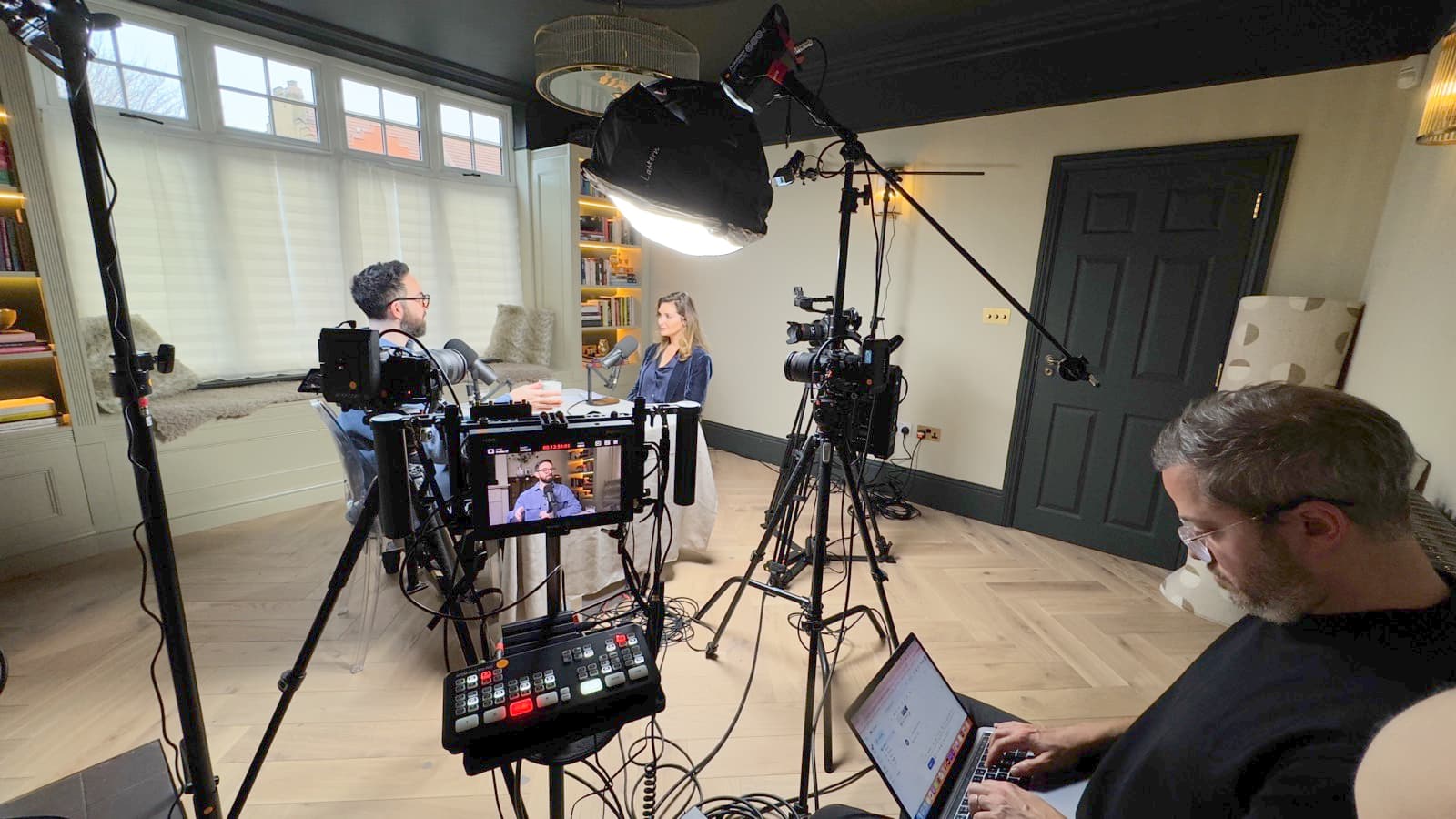 A panelled room mid-set-up, two cameras and a director's monitor, a producer working from a laptop in the foreground