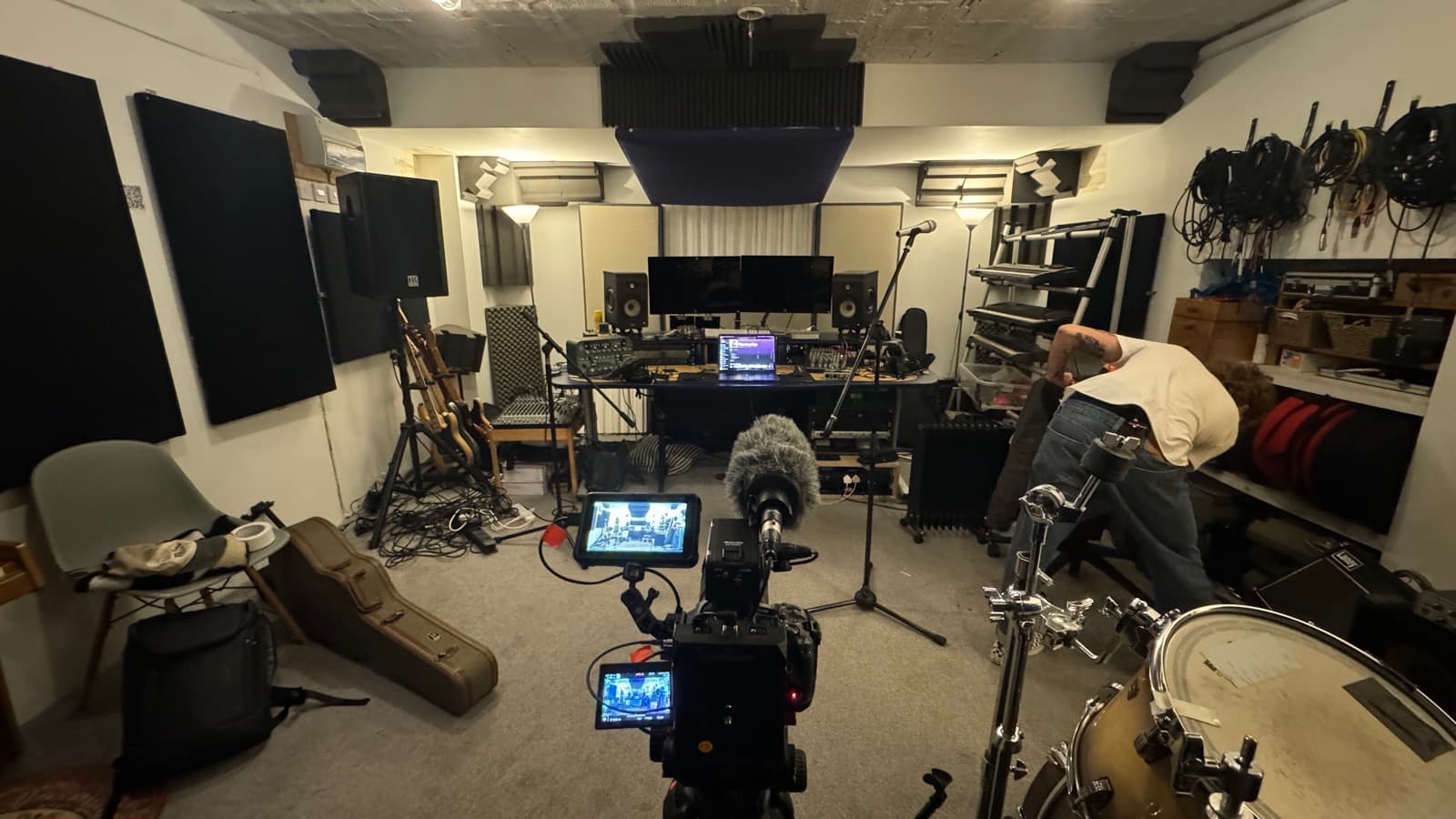 A rehearsal room with guitars on the wall and a kit in the corner, camera and shotgun mic set up in the foreground