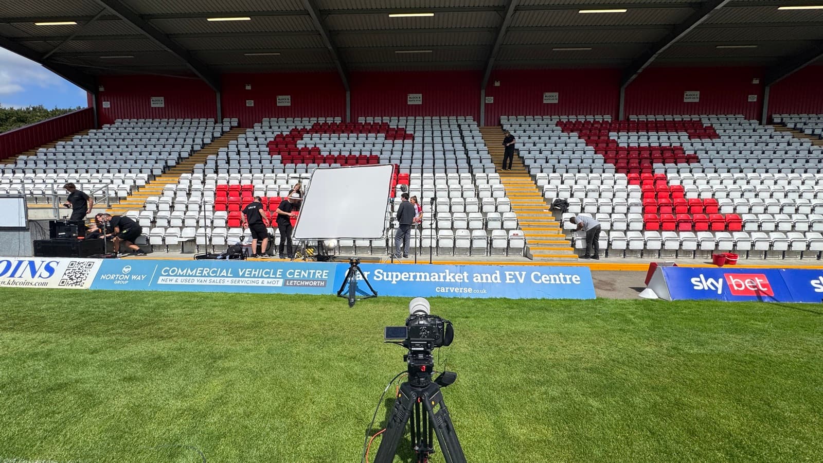 A camera on the pitch at a football ground, empty stand behind and a bounce board rigged across on the far touchline