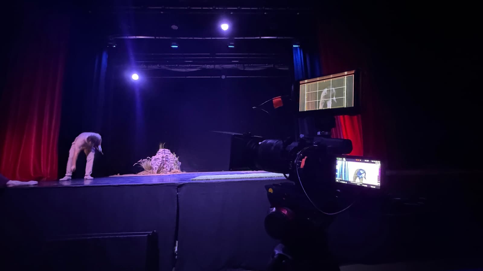 Covering a theatre stage lit blue against red curtains, camera and monitor at the lip of the stage