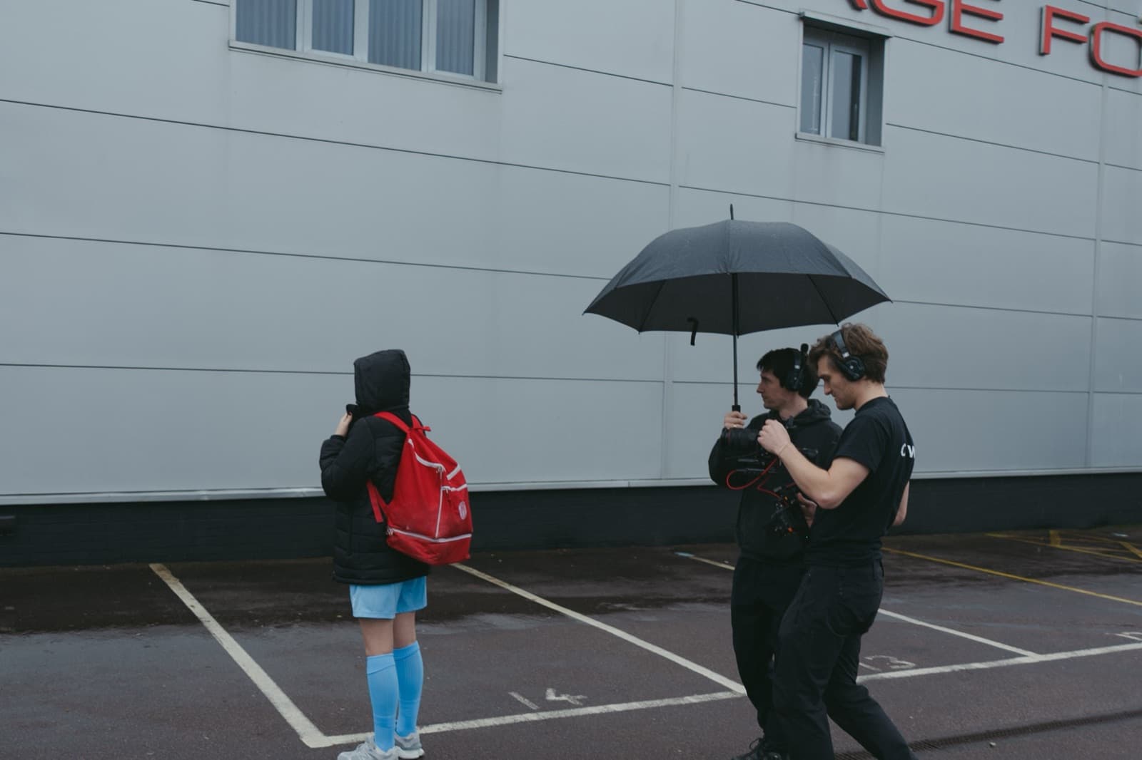 A contributor in kit waiting on a wet car park while the crew move in under an umbrella