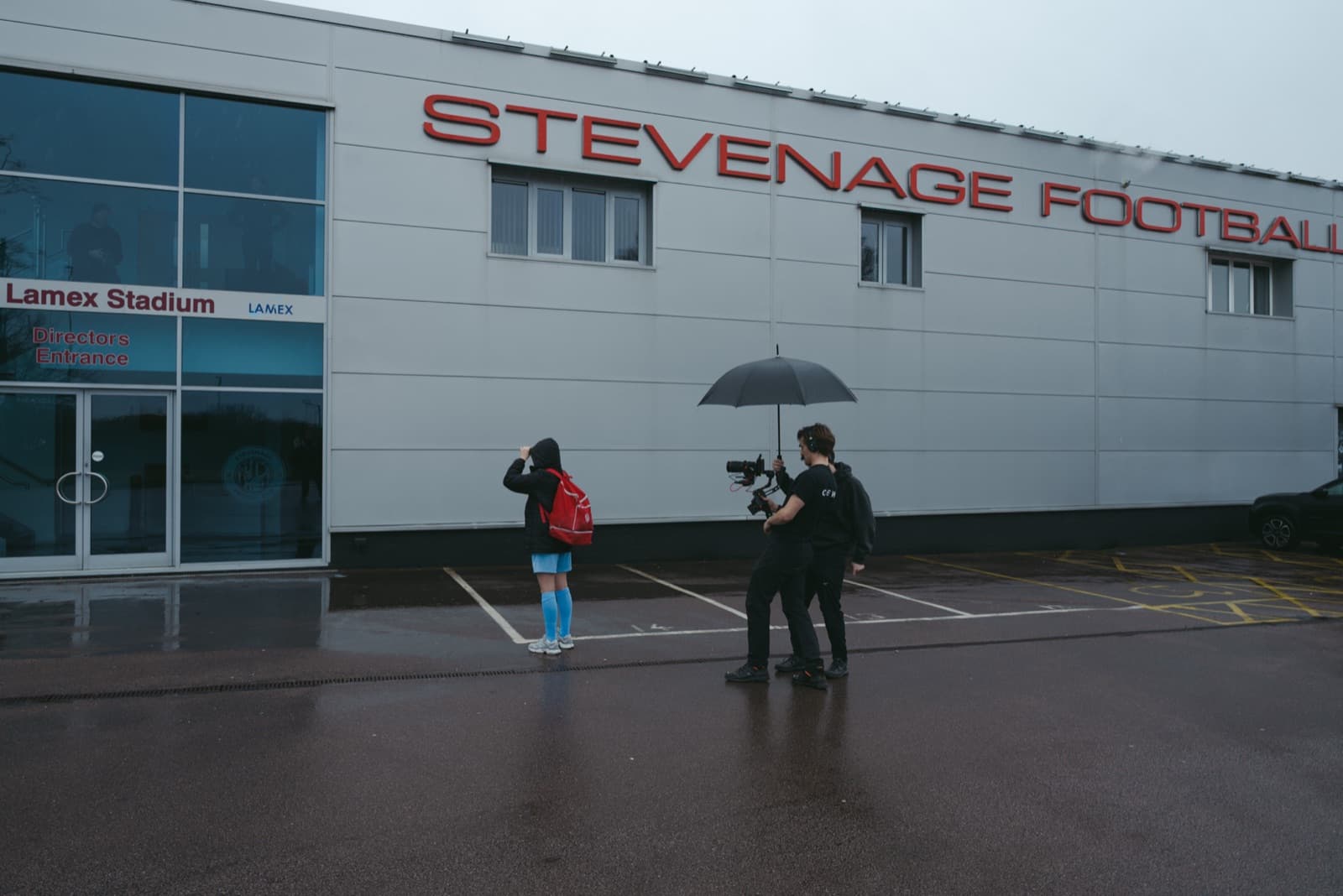 The stadium frontage in the rain, a contributor in kit in position and the crew moving in under an umbrella