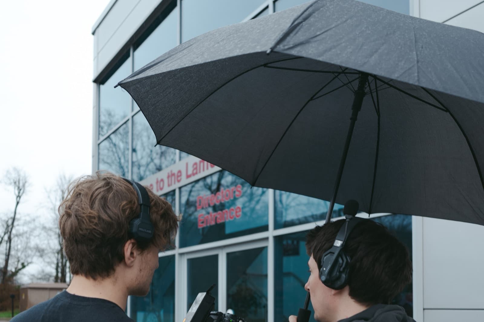 Two of the crew working under an umbrella in the rain, both on headphones at the camera